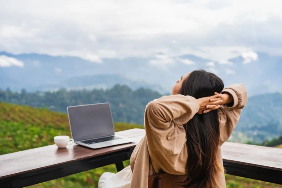 Frau sitzt entspannt auf einer Terrasse mit Bergblick, Laptop und Kaffee auf dem Tisch